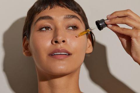 Close-up of woman applying facial oil with a dropper to her cheek, highlighting skin texture and natural lighting.