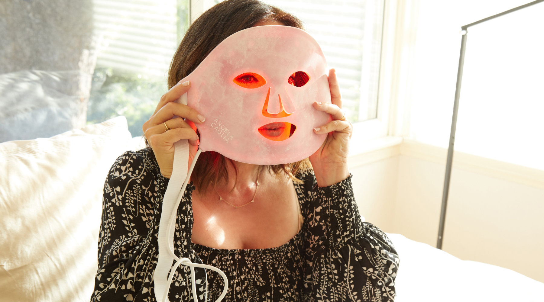 Woman holding pink ANGELA CAGLIA LED light-therapy facial mask with red lights glowing over her face